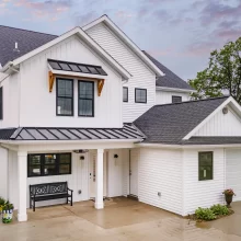 White lakefront home with black metal roof accents, covered front porch, and attached garage, lake visible in background