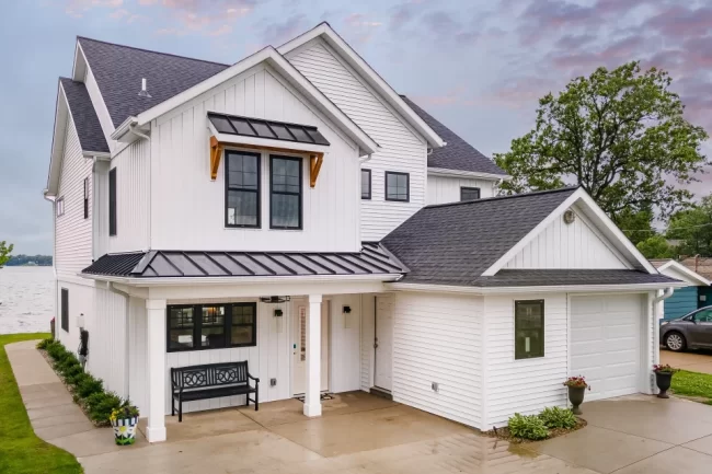 Lakefront Home Exterior with Covered Porch | Pennings & Sons White lakefront home with black metal roof accents, covered front porch, and attached garage, lake visible in background