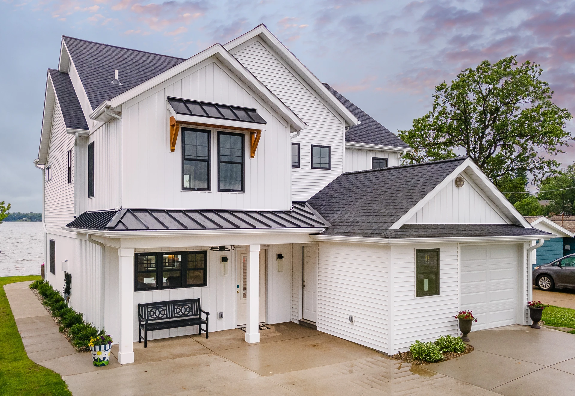White lakefront home with black metal roof accents, covered front porch, and attached garage, lake visible in background