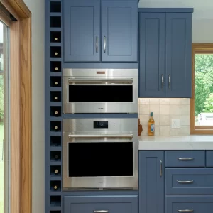 Blue kitchen cabinetry with built-in wine storage, stainless steel double ovens, white countertop, wood flooring, and natural wood window trim