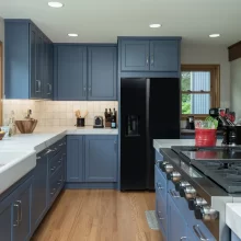 Remodeled kitchen with blue cabinets, farmhouse sink, black refrigerator, island cooktop, white countertops, and wood flooring