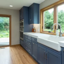 Blue kitchen cabinets with farmhouse sink, white countertops, wood window trim, stainless appliances, and views to the backyard deck