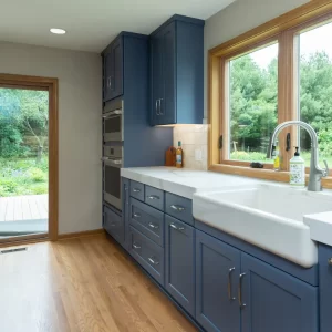 Blue kitchen cabinets with farmhouse sink, white countertops, wood window trim, stainless appliances, and views to the backyard deck
