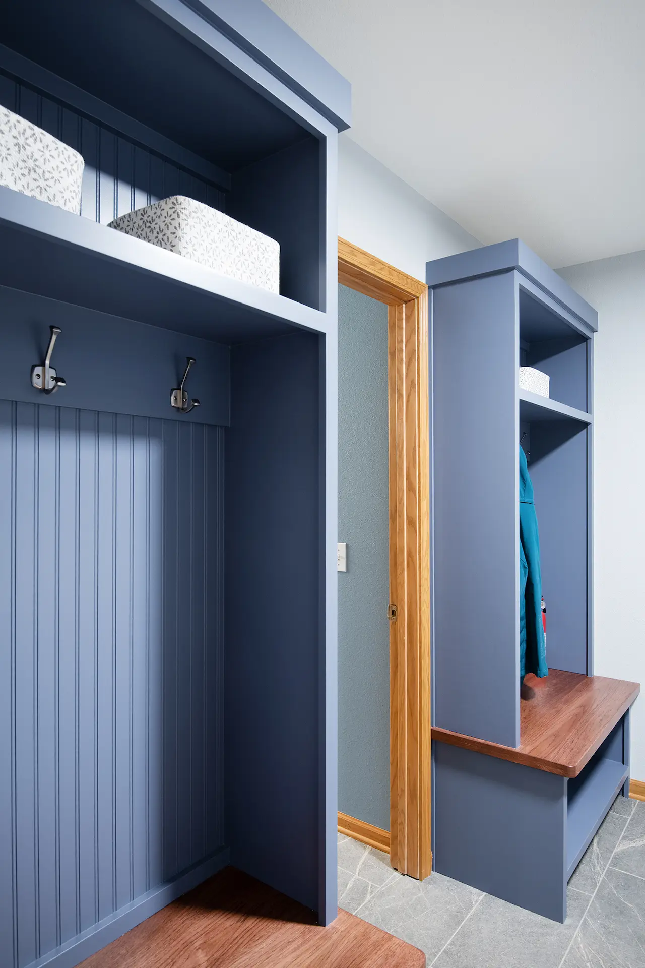 Custom blue mudroom lockers with wood bench seats, beadboard backing, coat hooks, upper storage shelves, and gray tile flooring