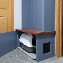 Custom blue cabinet with open door showing hidden litter box storage, wood bench seat, side pet entry, and gray tile flooring