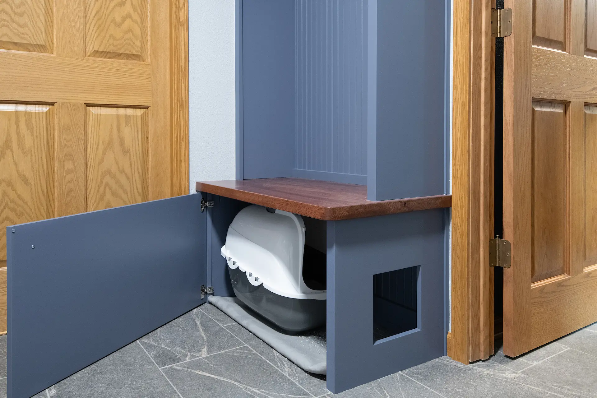 Custom blue cabinet with open door showing hidden litter box storage, wood bench seat, side pet entry, and gray tile flooring