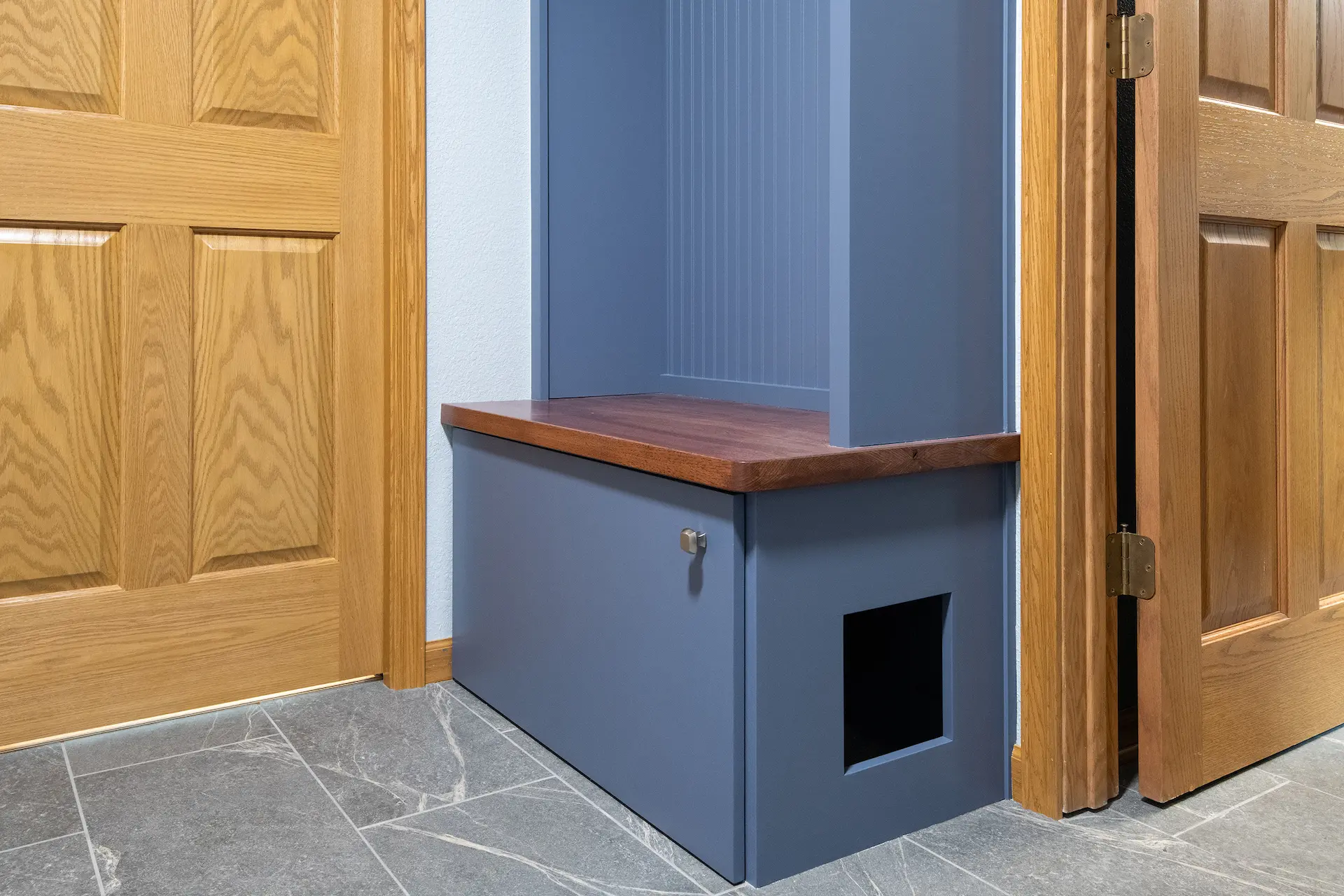 Custom blue mudroom bench with wood seat, cabinet storage, beadboard backing, gray tile floor, and built-in litter box opening