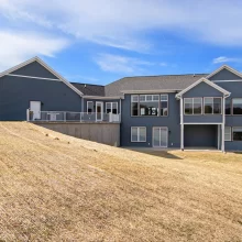 Rear view of a blue Galesburg custom home with large windows, walkout lower level, raised deck and sloped backyard.