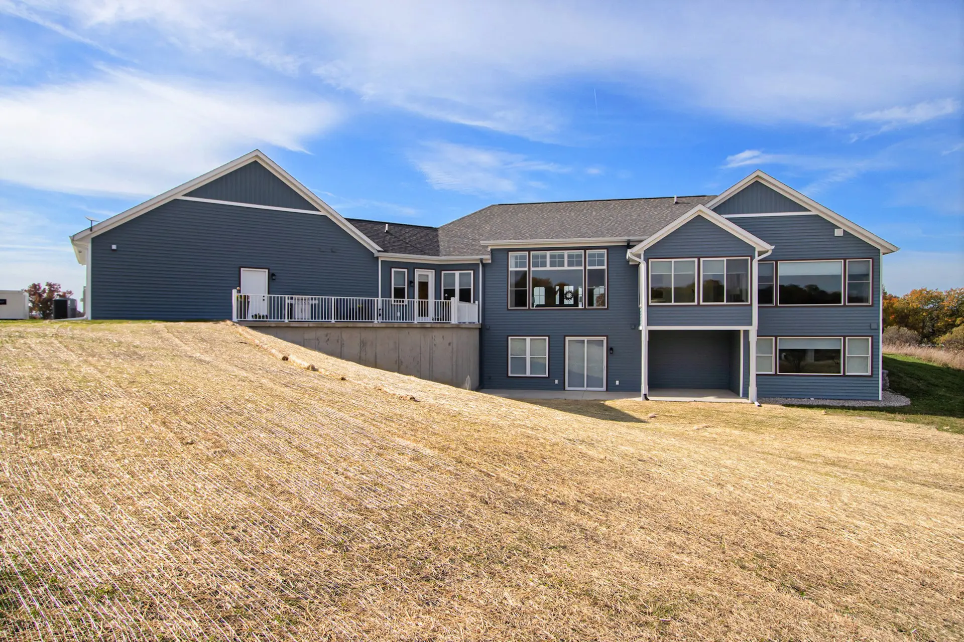 Rear view of a blue Galesburg custom home with large windows, walkout lower level, raised deck and sloped backyard.
