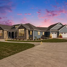 Twilight exterior view of a Galesburg custom home with warm interior lights, stone accents, blue siding and large driveway.