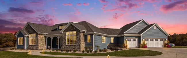 Galesburg Custom Home Exterior at Twilight | Pennings & Sons Twilight exterior view of a Galesburg custom home with warm interior lights, stone accents, blue siding and large driveway.