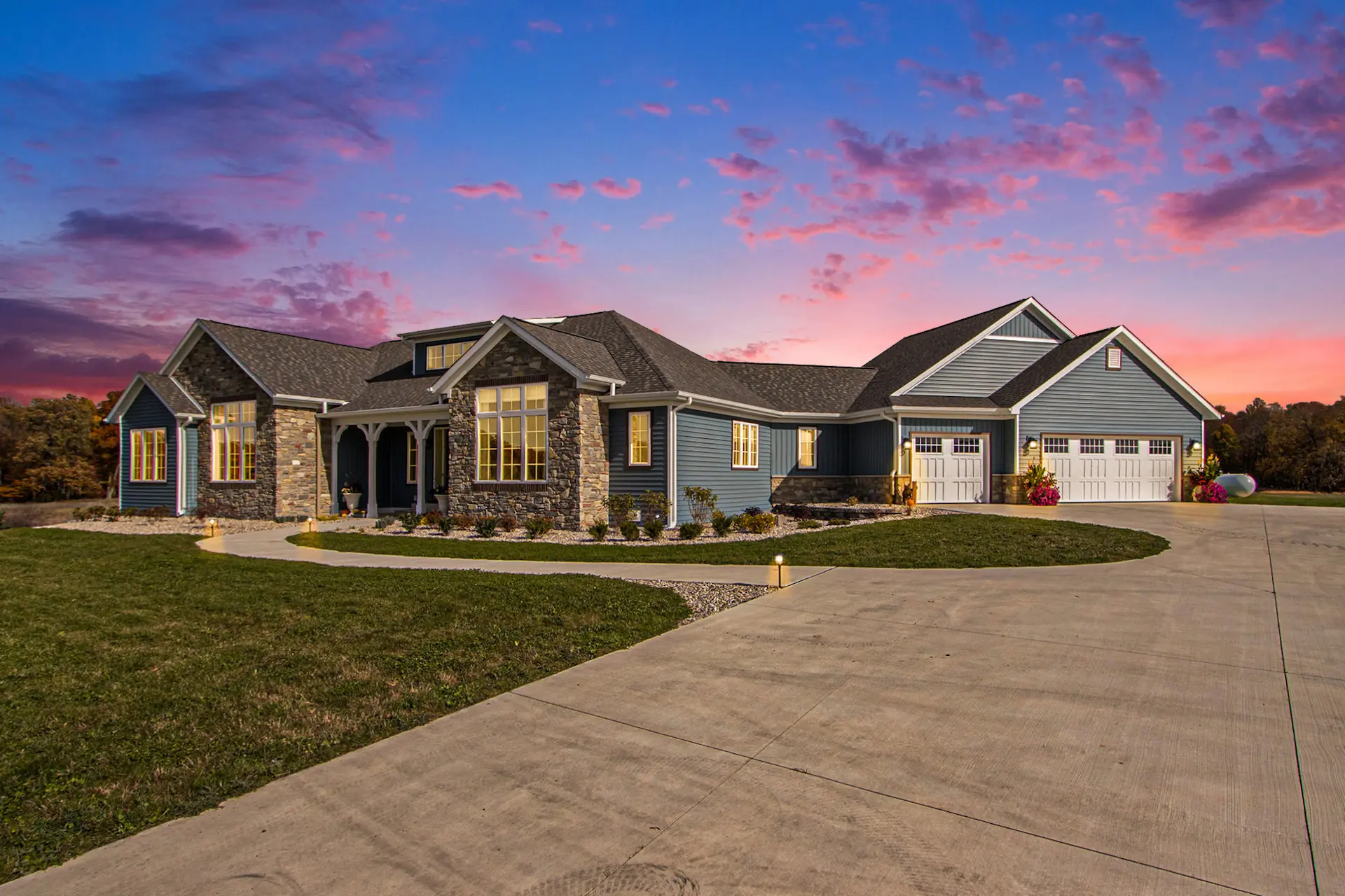 Twilight exterior view of a Galesburg custom home with warm interior lights, stone accents, blue siding and large driveway.