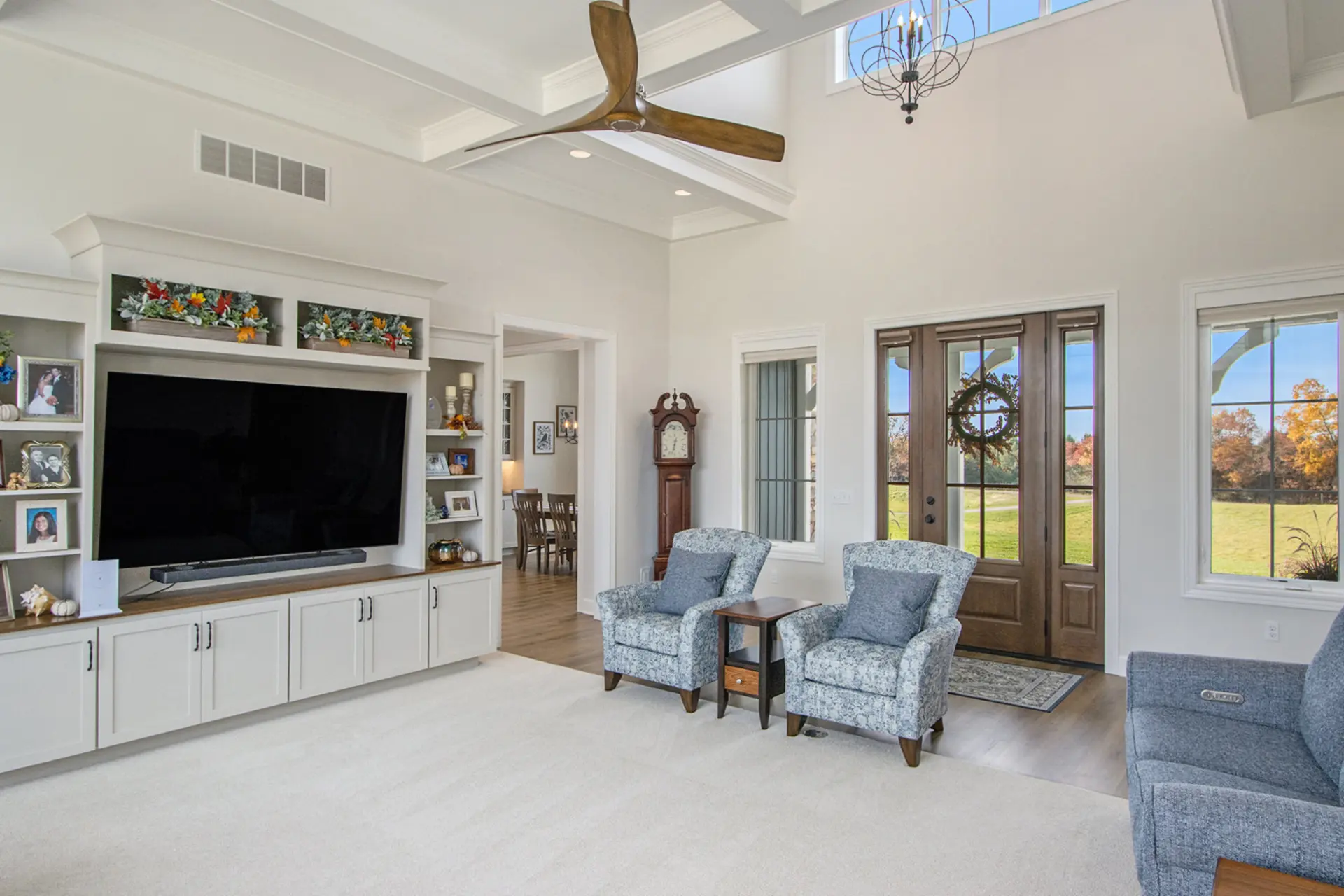 Living room with built-in media wall, blue accent chairs, vaulted ceiling, ceiling fan and front entry with wood doors.