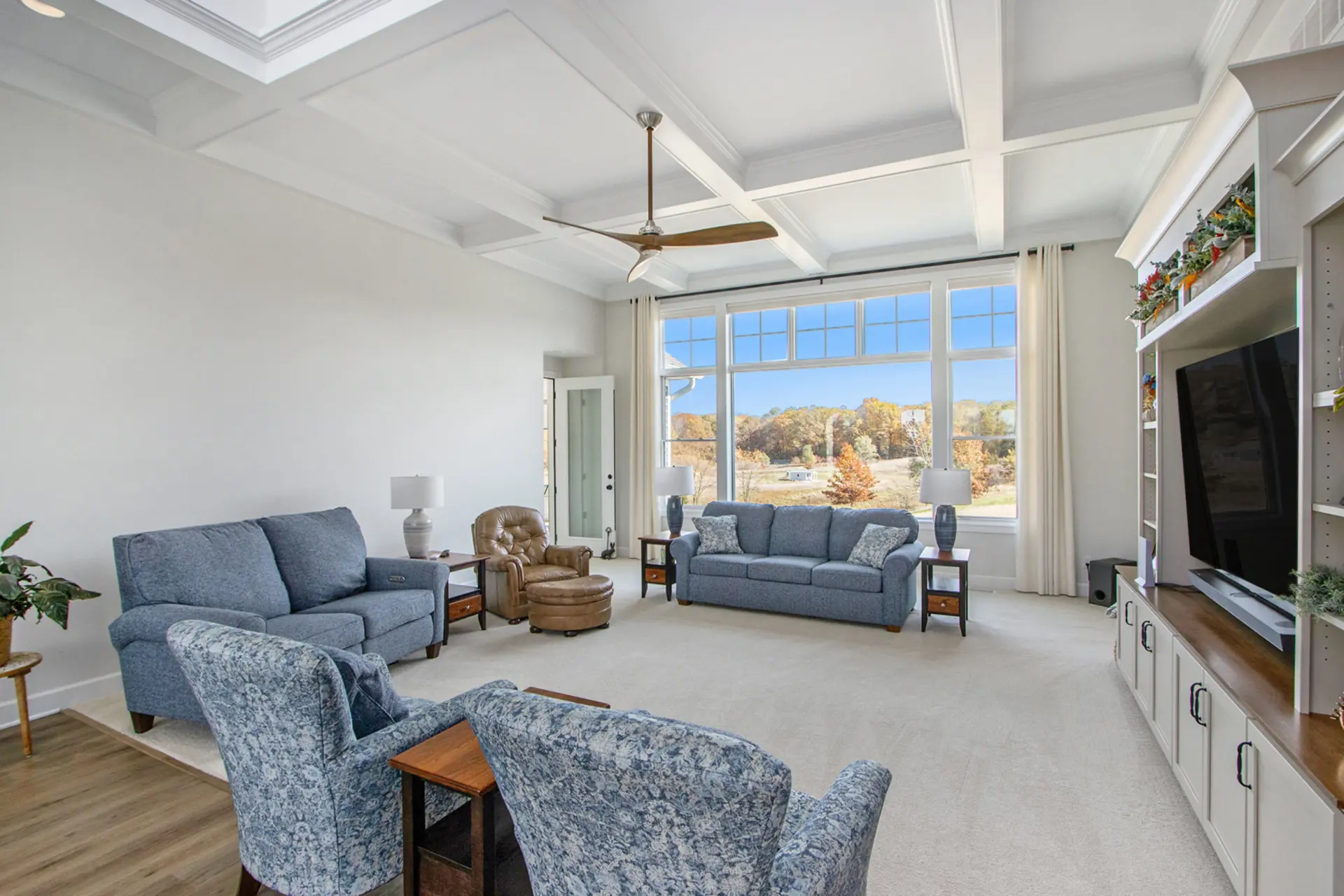 Bright living room in a Galesburg custom home with coffered ceiling, large windows, blue seating, built-in media wall and countryside views.