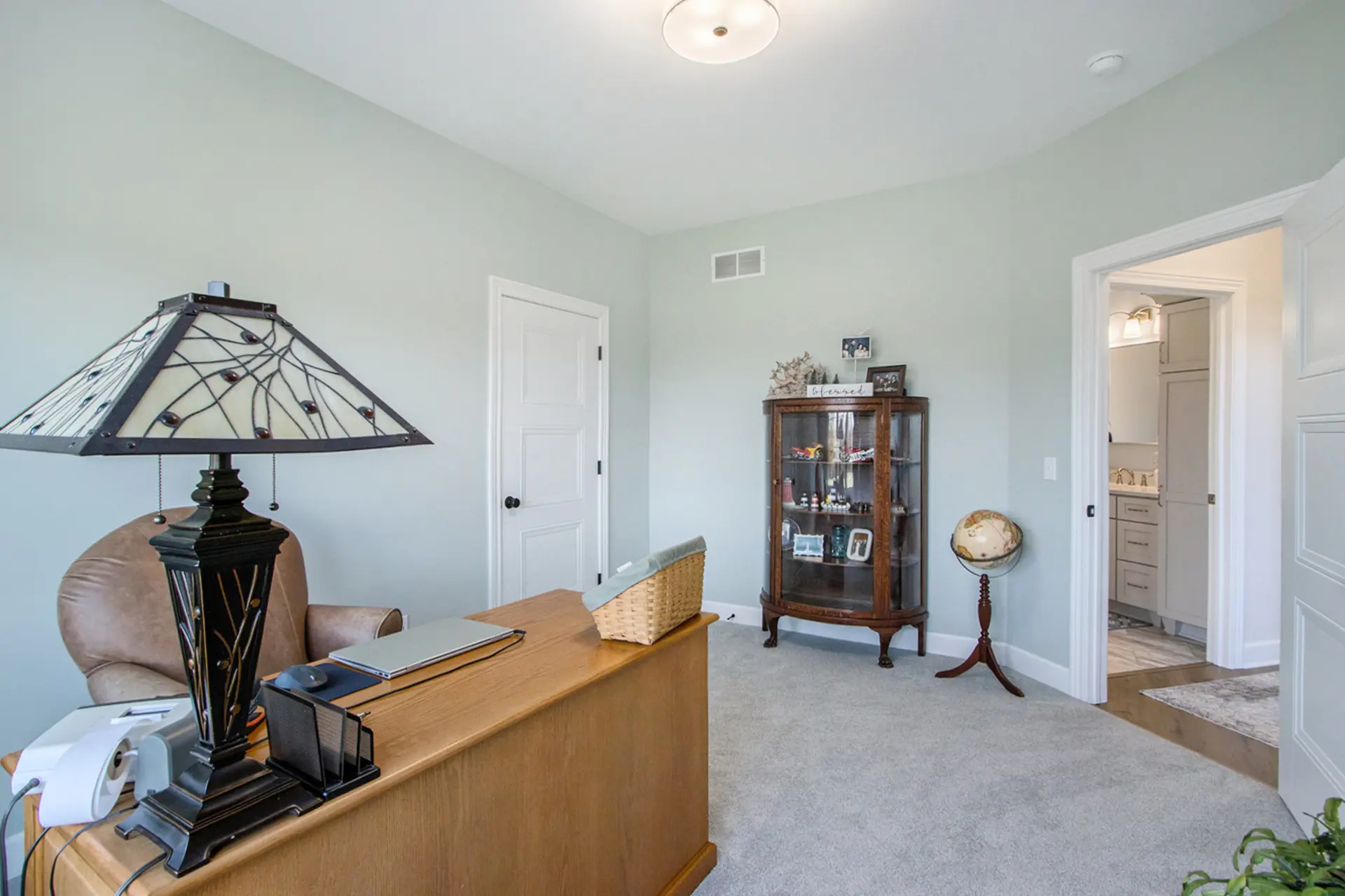 Home office angle showing wood desk, display cabinet, globe, soft green walls and doorway to a nearby bathroom.
