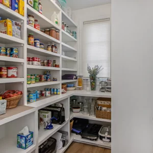 Walk-in pantry with open white shelving, organized food storage, small window and built-in countertop space.