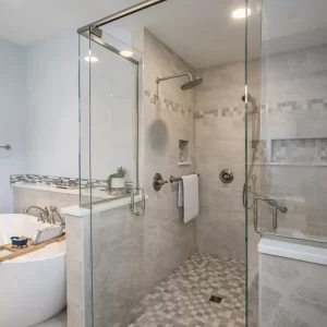 Glass-enclosed primary bathroom shower with neutral wall tile, mosaic floor tile, built-in niche and nearby soaking tub.