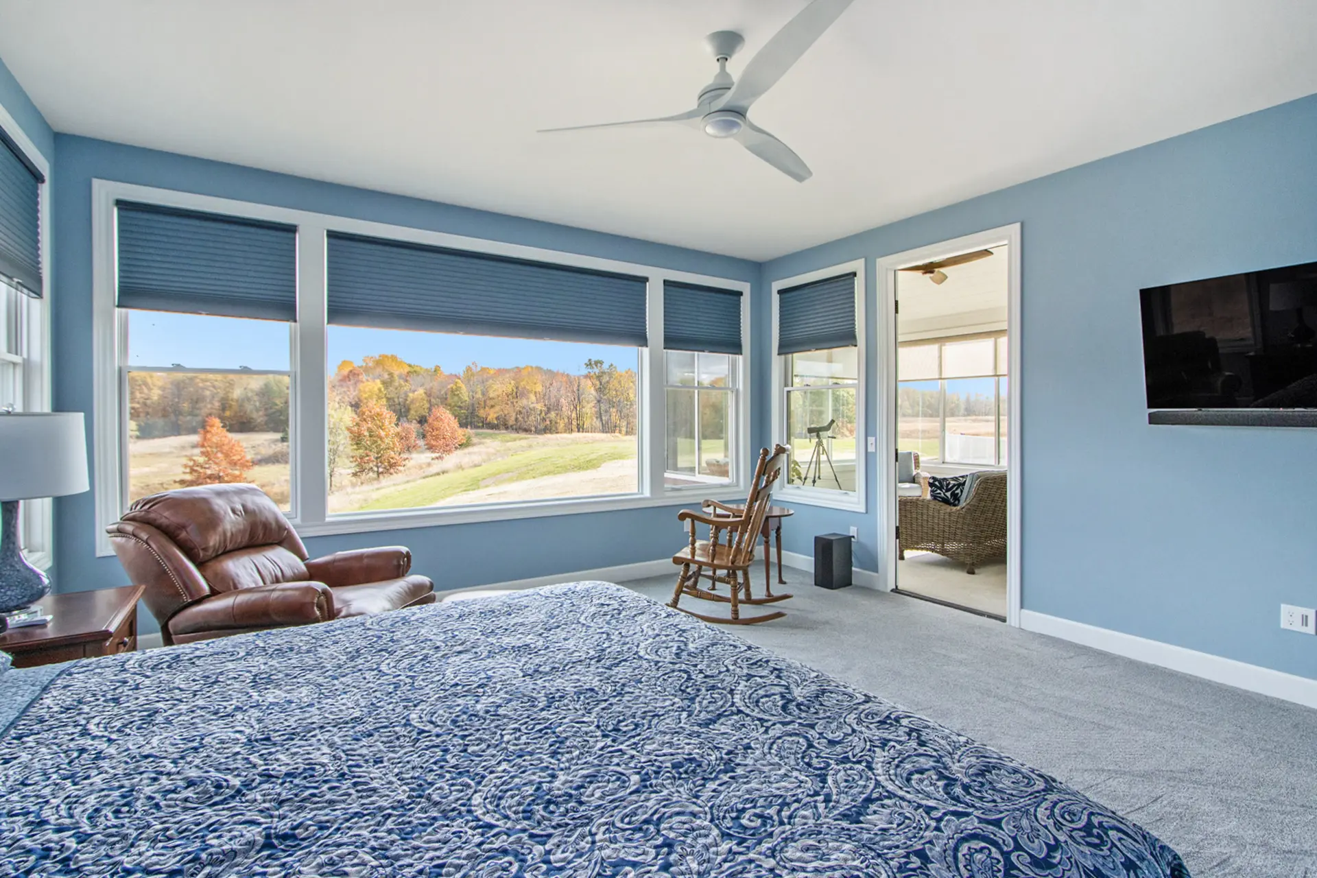Primary bedroom with blue walls, large windows, countryside views, ceiling fan, sitting area and access to a screened porch.
