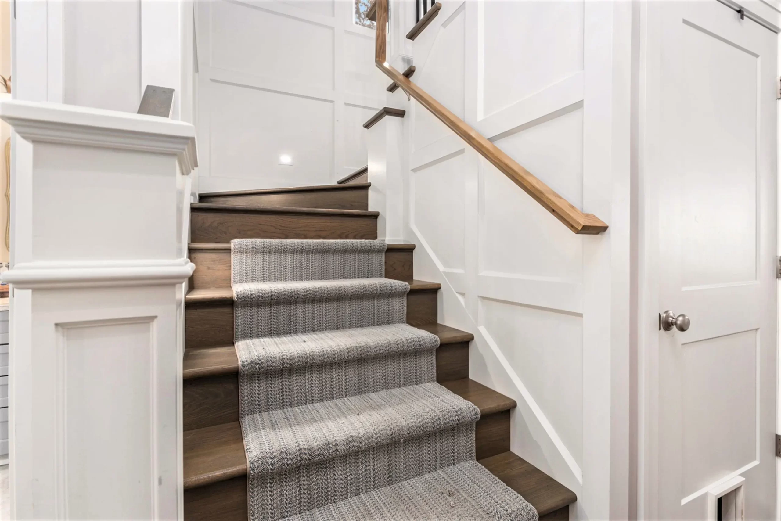 Close-up of custom staircase with wood treads, textured carpet runner, borad and batten wall pattern, and warm wood handrail.