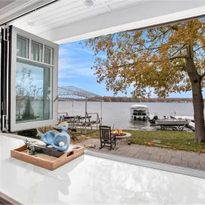 Open folding kitchen window framing a lakefront patio, fire pit area, dock, boat lift, and mature waterfront tree