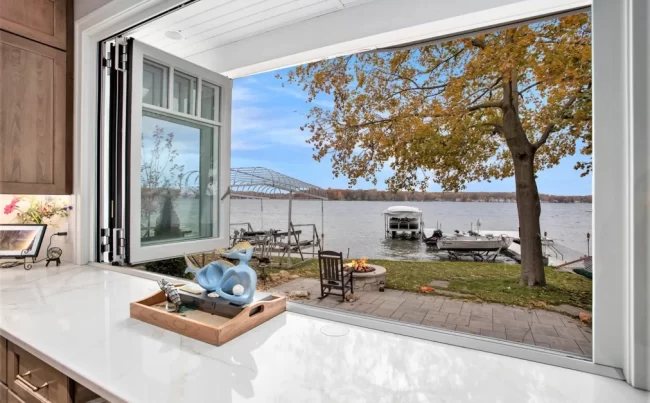 Kitchen Pass-Through Window to Lakefront Patio | Pennings & Sons Open folding kitchen window framing a lakefront patio, fire pit area, dock, boat lift, and mature waterfront tree