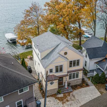 Aerial view of a lakefront custom home with waterfront lawn, dock access, mature trees, and neighboring cottages