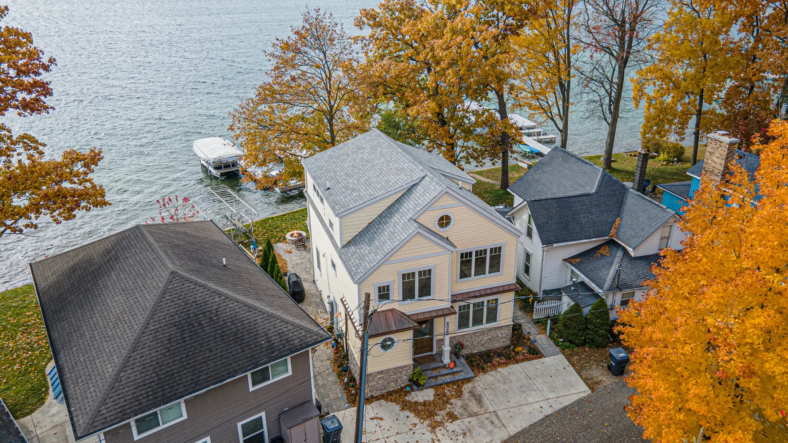 Aerial view of a lakefront custom home with waterfront lawn, dock access, mature trees, and neighboring cottages