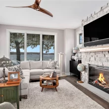 Living room with large lake-facing windows, stone fireplace, mounted TV, neutral seating, and hardwood flooring