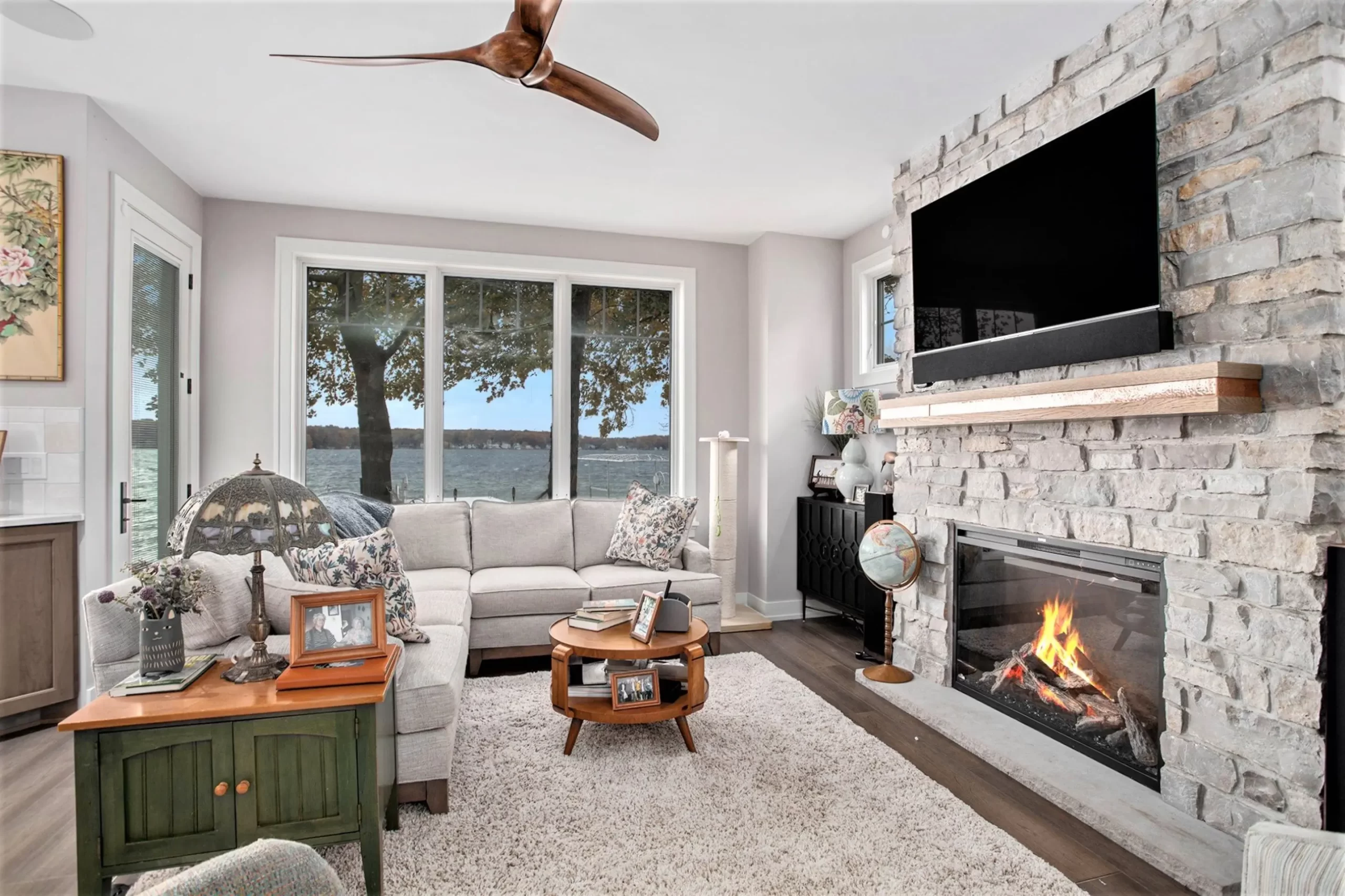 Living room with large lake-facing windows, stone fireplace, mounted TV, neutral seating, and hardwood flooring