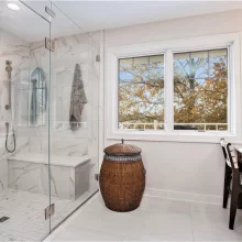 Primary bathroom with frameless glass shower, marble-look wall tile, built-in bench, lake view window, and wood storage