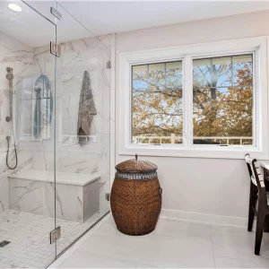 Primary bathroom with frameless glass shower, marble-look wall tile, built-in bench, lake view window, and wood storage