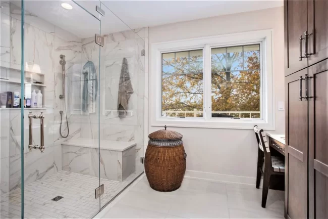 Primary Bath Glass Shower with Lake View Window | Pennings & Sons Primary bathroom with frameless glass shower, marble-look wall tile, built-in bench, lake view window, and wood storage