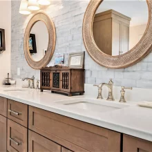 Close-up of primary bath wood vanity with white countertop, polished faucets, round mirrors, and marble-look brick lay backsplash tile