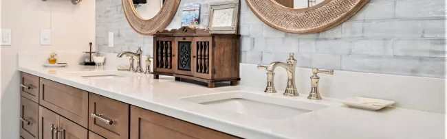 Close-up of primary bath wood vanity with white countertop, polished faucets, round mirrors, and marble-look brick lay backsplash tile