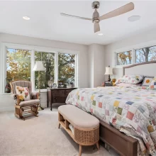 Primary bedroom with large windows, neutral carpet, wood furniture, ceiling fan, upholstered bed, and colorful quilt