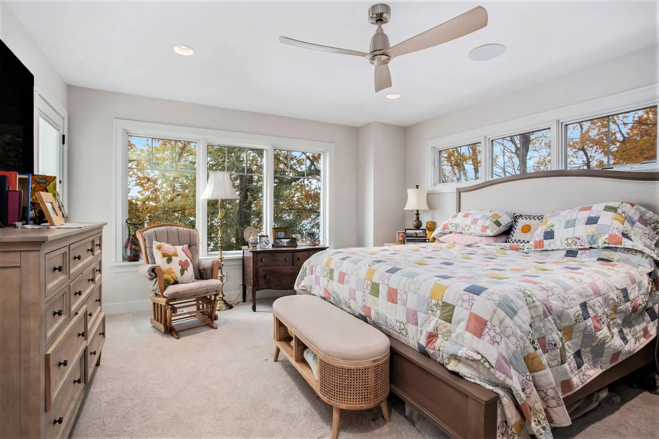 Primary bedroom with large windows, neutral carpet, wood furniture, ceiling fan, upholstered bed, and colorful quilt