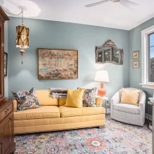 Cozy sitting room with blue walls, yellow sofa, patterned rug, vintage hutch, artwork, and bright window seating