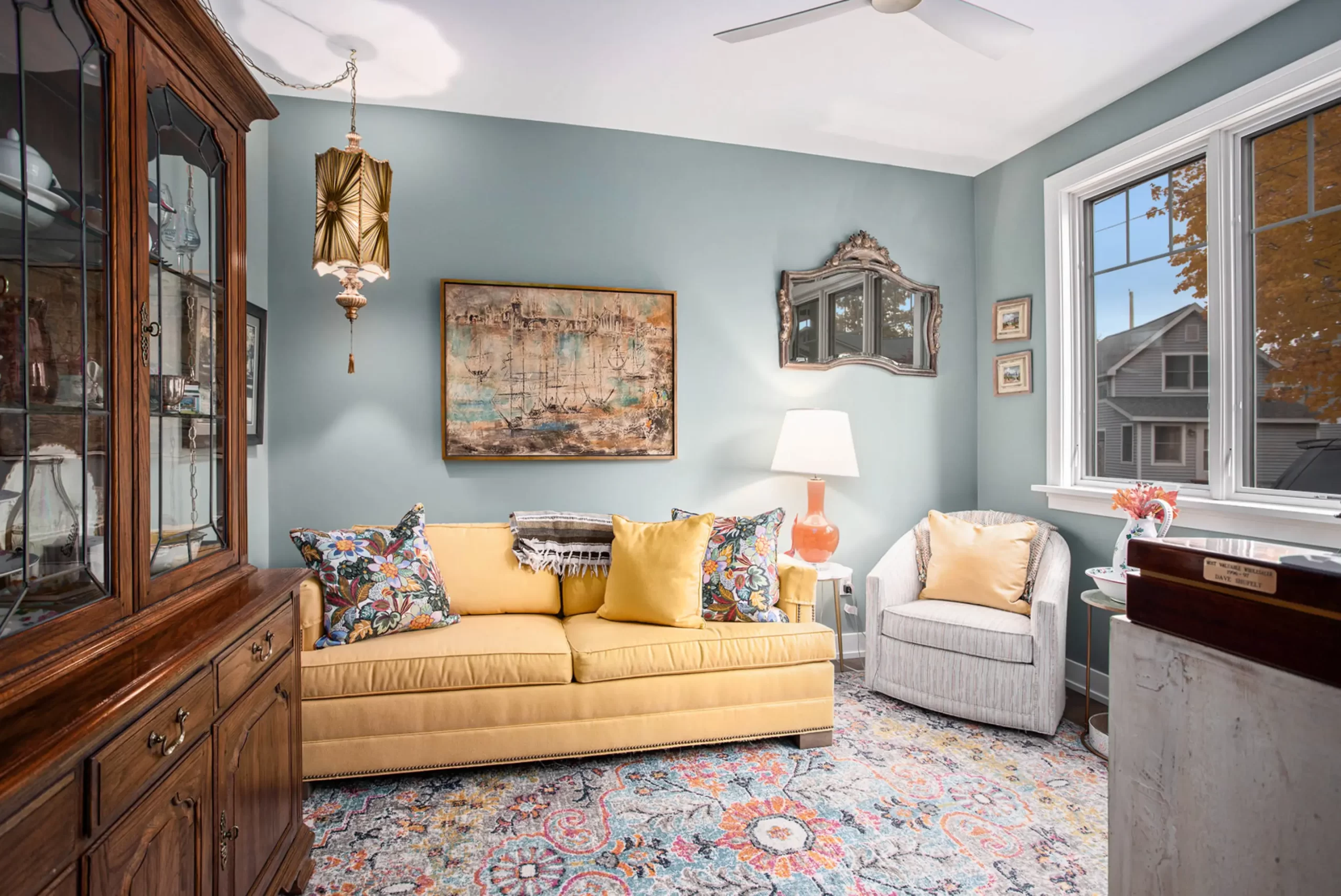 Cozy sitting room with blue walls, yellow sofa, patterned rug, vintage hutch, artwork, and bright window seating
