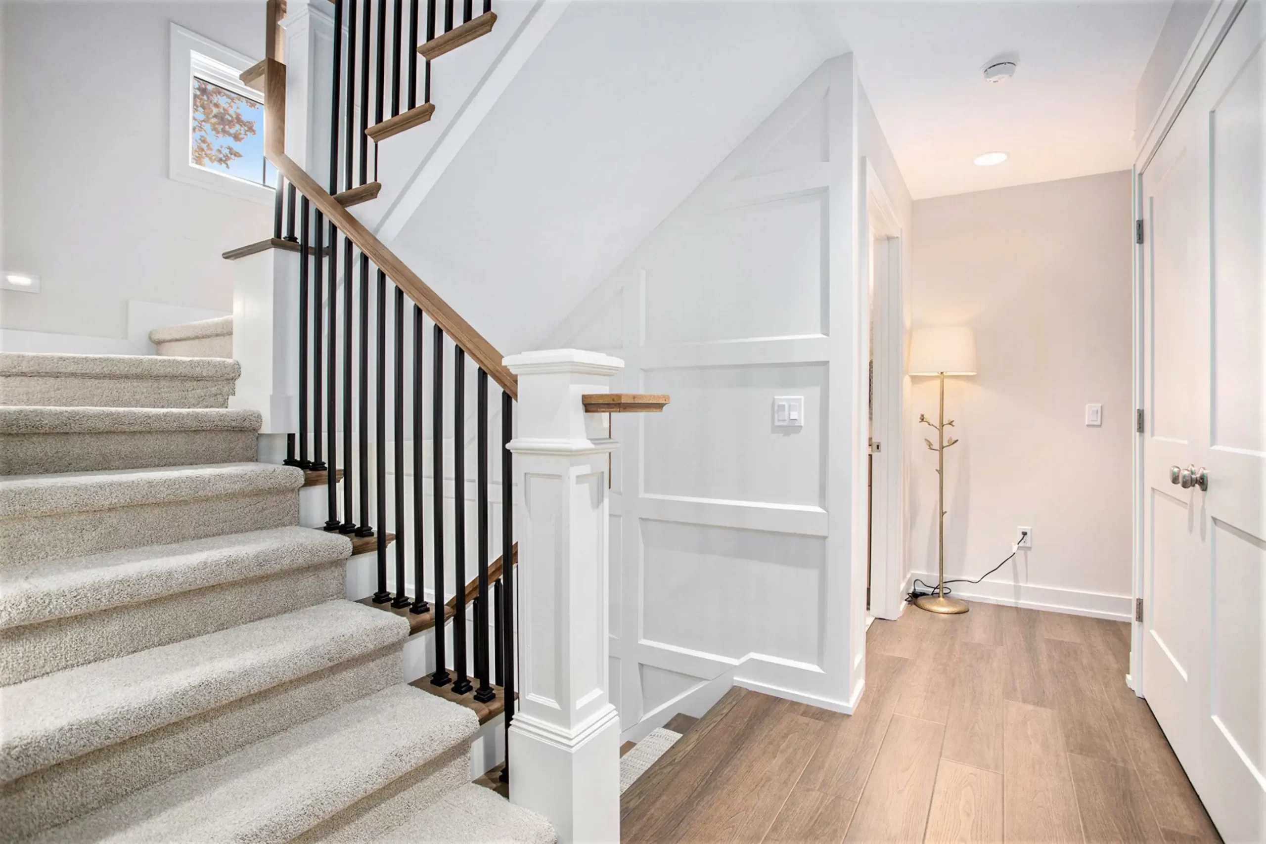 Stair landing with white borad and batten wall pattern, black balusters, wood handrail, hardwood flooring, and bright hallway lighting