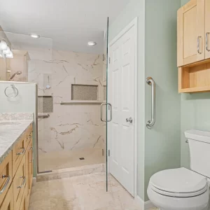 Accessible bathroom remodel with walk-in shower, glass door, grab bars, double vanity, and light wood cabinetry