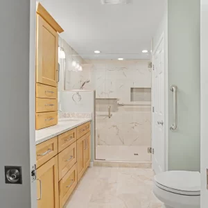 Bathroom entry view showing custom vanity cabinets, walk-in shower, glass door, toilet, and safety grab bar