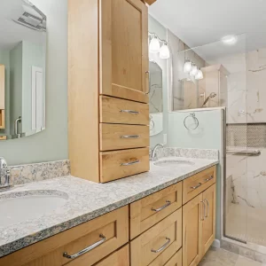 Custom bathroom vanity with light wood cabinets, granite-style countertop, mirrors, and accessible tiled shower