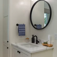Bathroom vanity with white cabinetry, black faucet, round black mirror, glass vanity lights, blue hand towel, and tall storage cabinet