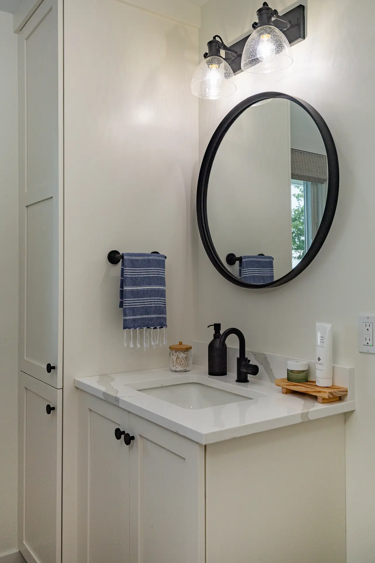 Bathroom vanity with white cabinetry, black faucet, round black mirror, glass vanity lights, blue hand towel, and tall storage cabinet