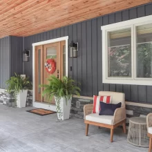 Covered porch entry with wood front door, blue vertical siding, stone accents, wood ceiling, outdoor chairs, and potted ferns