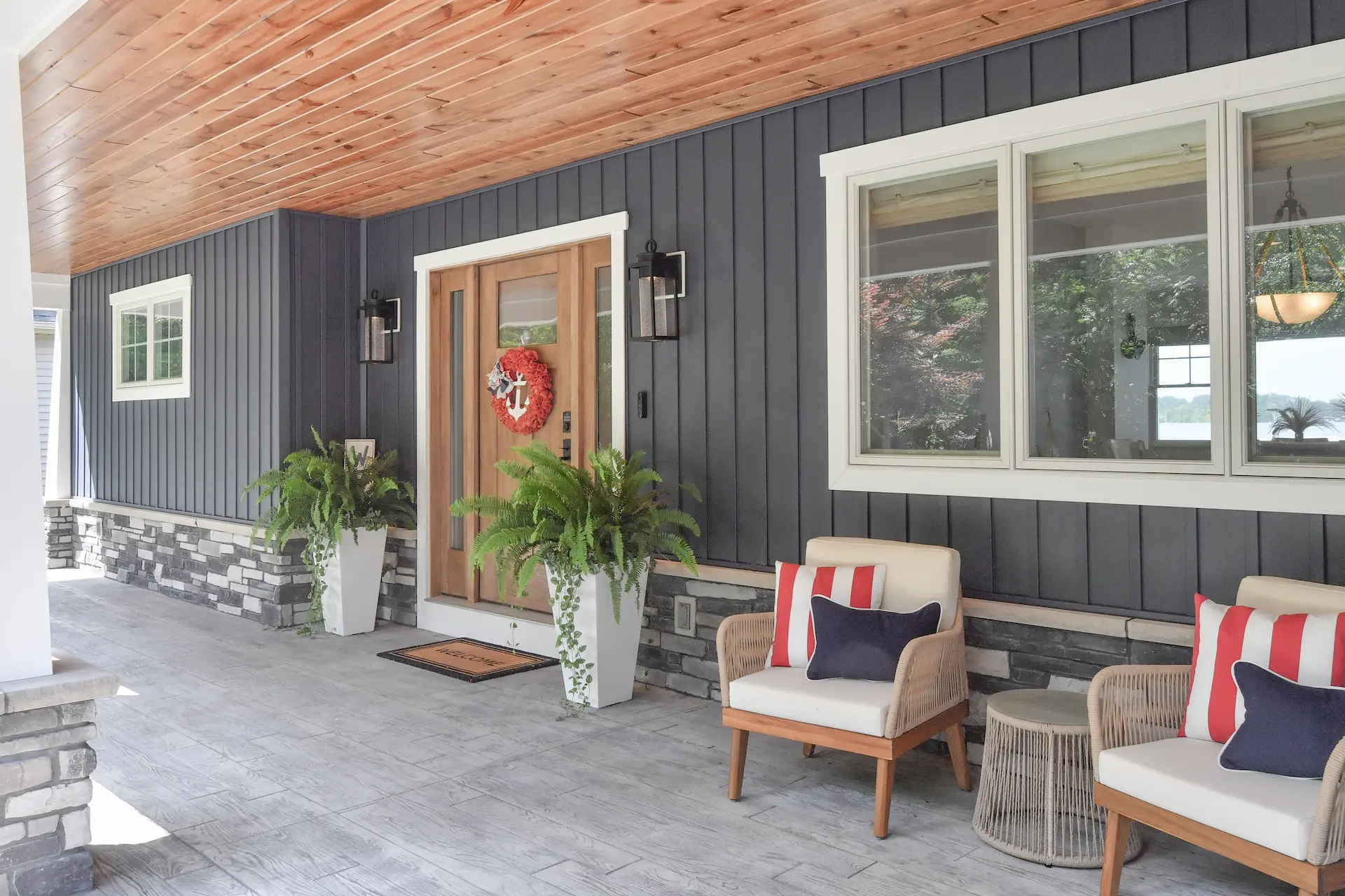 Covered porch entry with wood front door, blue vertical siding, stone accents, wood ceiling, outdoor chairs, and potted ferns