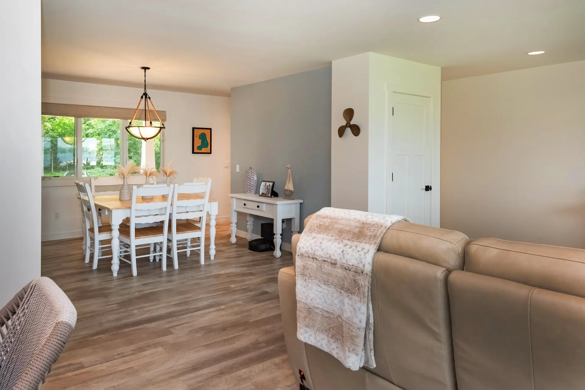 Open dining and living area with white dining table, pendant light, neutral seating, wood-look flooring, and bright natural light