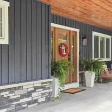 Front porch with wood ceiling, blue vertical siding, stone base, wood entry door, outdoor seating, potted ferns, and white trim