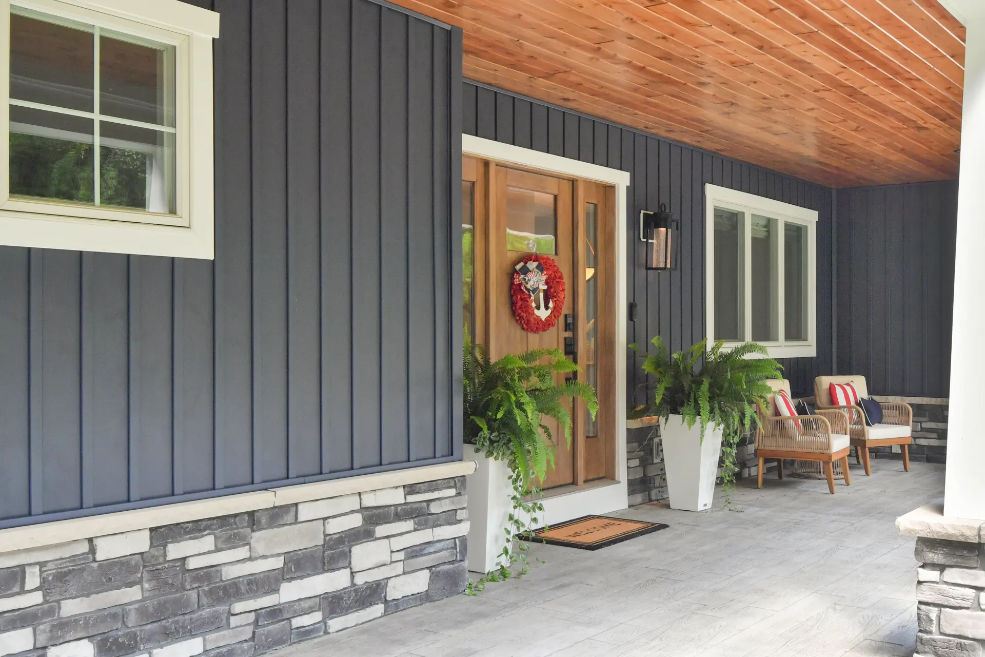 Front porch with wood ceiling, blue vertical siding, stone base, wood entry door, outdoor seating, potted ferns, and white trim