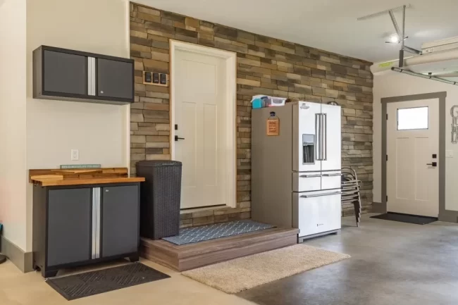 Finished garage entry area with stone accent wall, raised platform, exterior door, storage cabinets, refrigerator, and concrete floor